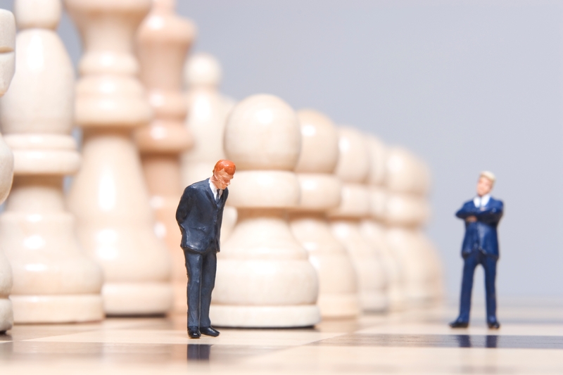 Two miniature figurines of men in suits stand on a chessboard, surrounded by oversized chess pieces. One man appears pensive with his head lowered, contemplating public policy, while the other stands confidently with arms crossed. The background is a blurred array of chess pawns.