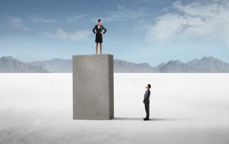 A businesswoman stands confidently on top of a tall concrete block, looking down at a businessman who stands on the ground, gazing up at her. Set against a vast, desolate landscape with mountains in the background and a blue sky overhead, this scene highlights women's rising influence in the corporate world.
