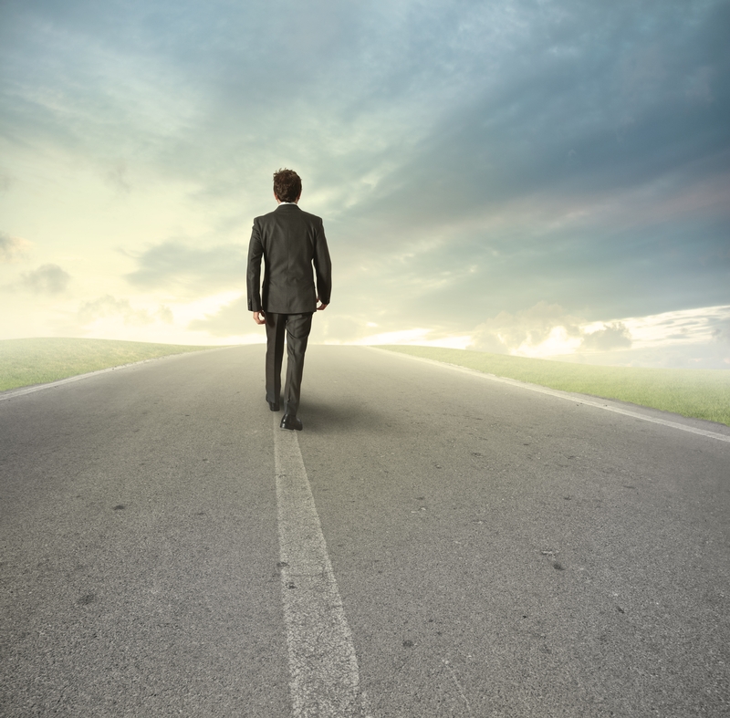 A leader in a suit walks alone on a deserted road stretching towards the horizon, under a dramatic sky with clouds. The scene suggests a journey or pathway, with the man seen from behind, moving towards a distant vanishing point.