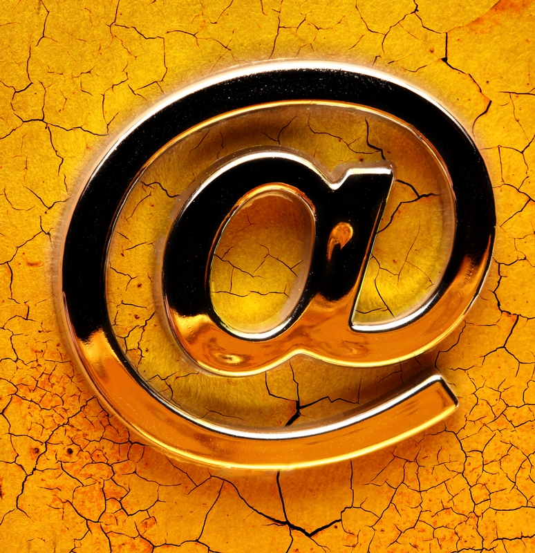 A shiny, metallic "@" symbol representing an email address is centered against a dry, cracked, yellow-orange earth background, creating a striking contrast between the smooth, reflective surface of the symbol and the rough, fragmented texture of the ground.