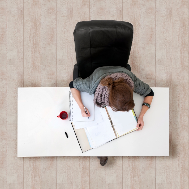 A person with brown hair is seated at a white desk, writing in a notebook and referencing an open binder filled with GRE prep materials. A red coffee cup and a black pen are also on the desk, which rests on a wooden floor. The photo is taken from an overhead perspective.