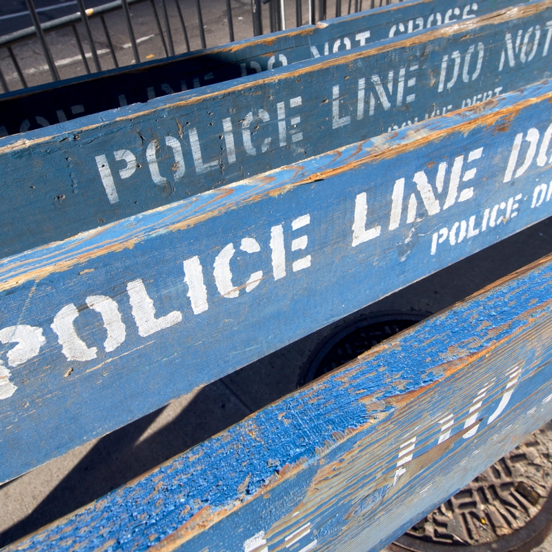 Blue wooden barriers with "POLICE LINE DO NOT CROSS" stenciled in white are stacked on the street. The barriers, weathered and peeling, reflect years of service under the watchful eye of the Police Chief. In the background, a metal fence and part of a manhole cover can be seen.