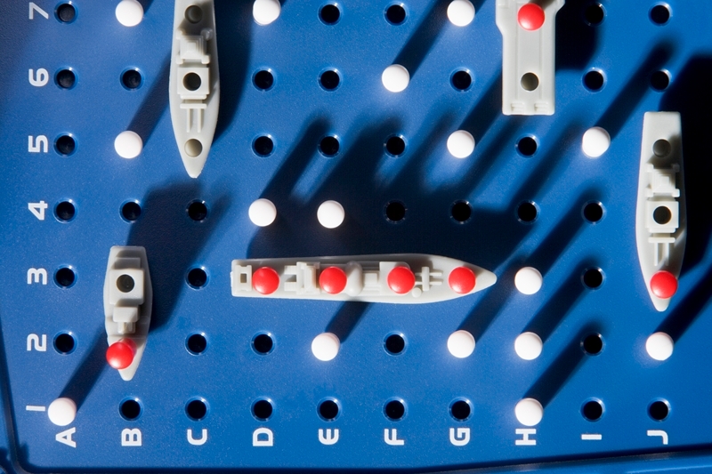 A close-up of a Battleship game board reveals an intense contest, reminiscent of international relations. Several ships with red pegs indicating hits are positioned on the blue, peg-filled grid. The grid is labeled with numbers 1-5 on the left and letters A-I on the bottom.