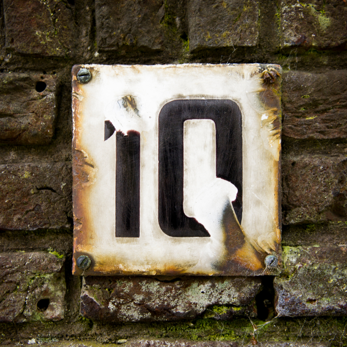 A weathered rectangular metal plate displaying the number "10" is mounted on a textured, moss-covered brick wall. The plate has rust and chipped paint, giving it an aged, rustic appearance akin to an old interview location. Four screws anchor the plate to the wall.