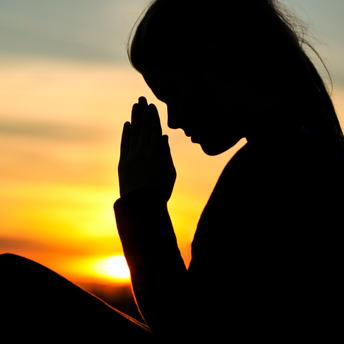 Silhouette of a person with long hair, facing sideways, holding their hands together in a prayer-like gesture. The background features a vibrant sunset with a warm orange and yellow sky, capturing an essence of humility.