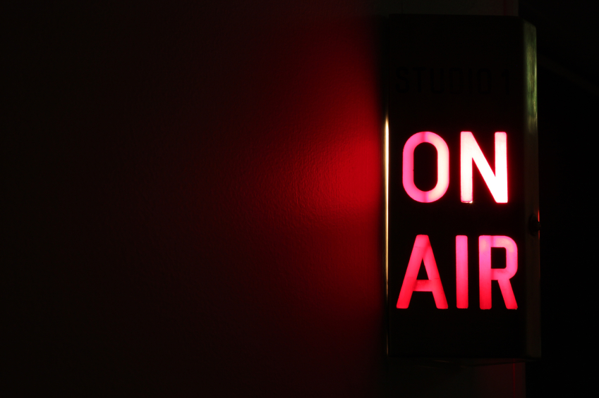 A red and white illuminated "ON AIR" sign is affixed to a dark wall, indicating that a live broadcast or recording is in progress in Studio 1. The room is dimly lit, with the sign casting a red glow on the nearby wall as if resuming an unspoken tradition.