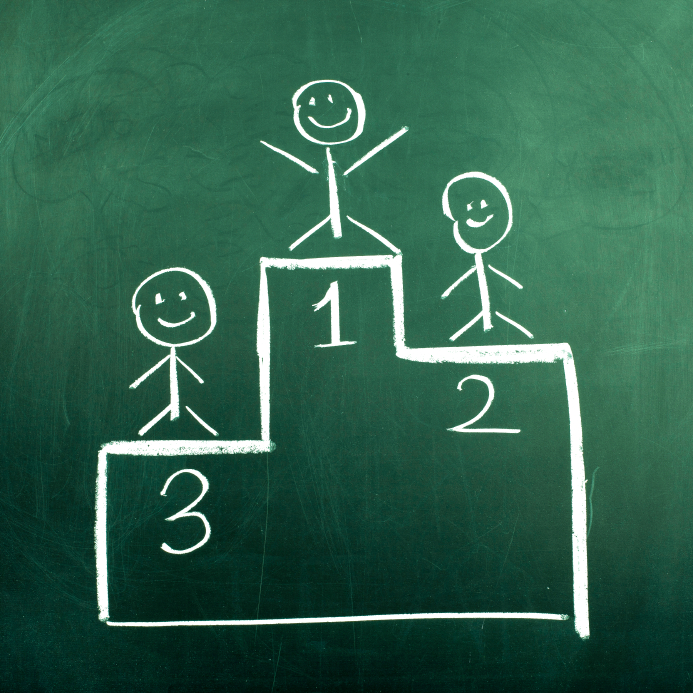 A chalk drawing on a green chalkboard depicts three stick figures on a winners' podium, with the top tier labeled "1" and arms raised in victory. The second and third tiers, labeled "2" and "3," each have a figure standing proudly to receive their award.