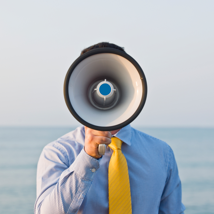 A candidate stands by the ocean, holding a white and black megaphone in front of their face, obscuring their identity. They are wearing a blue collared shirt with vertical stripes and a bright yellow tie. The sky and calm sea form the background.
