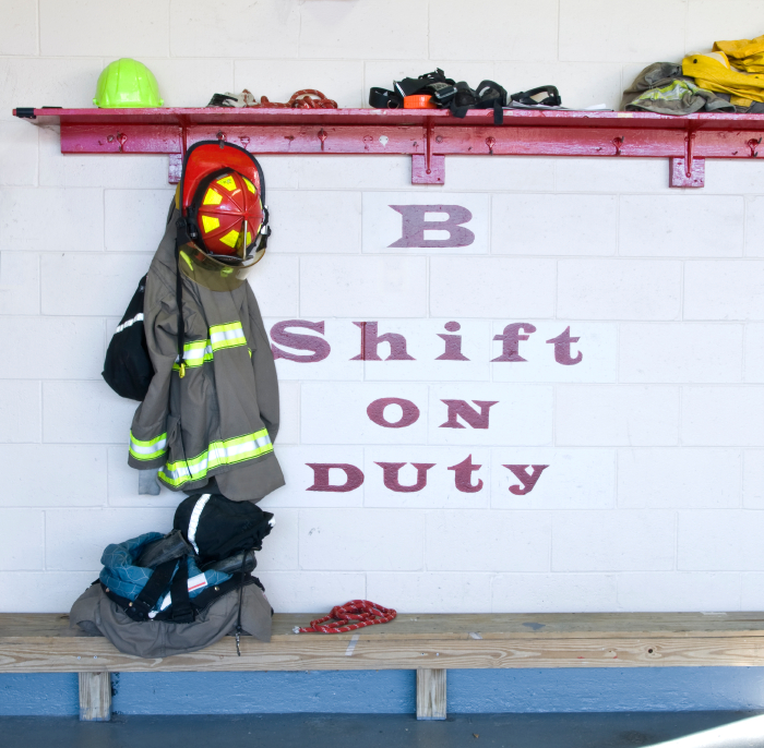 A wall with the text "B Shift on Duty" painted on it. Present above are firefighter gear including a helmet, jacket, and gloves. Additional items are stored on a red shelf, and more clothing rests on a bench below.