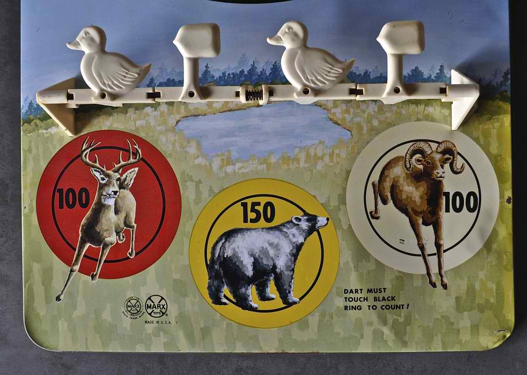 A vintage dart game board featuring illustrations of a deer on a red circle worth 100 points, a badger on a yellow circle worth 150 points, and a ram on a white circle worth 100 points. Ducks are mounted on a bar at the top for dart holding, making it as thrilling as job hunting with every throw.