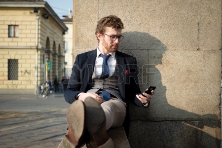 A man in a suit and tie, with glasses and short curly hair, is sitting against a stone wall, looking at his smartphone. He is outdoors in a sunny urban area, with buildings and a street visible in the background. He appears to be relaxed and focused on his phone, possibly checking local government updates.