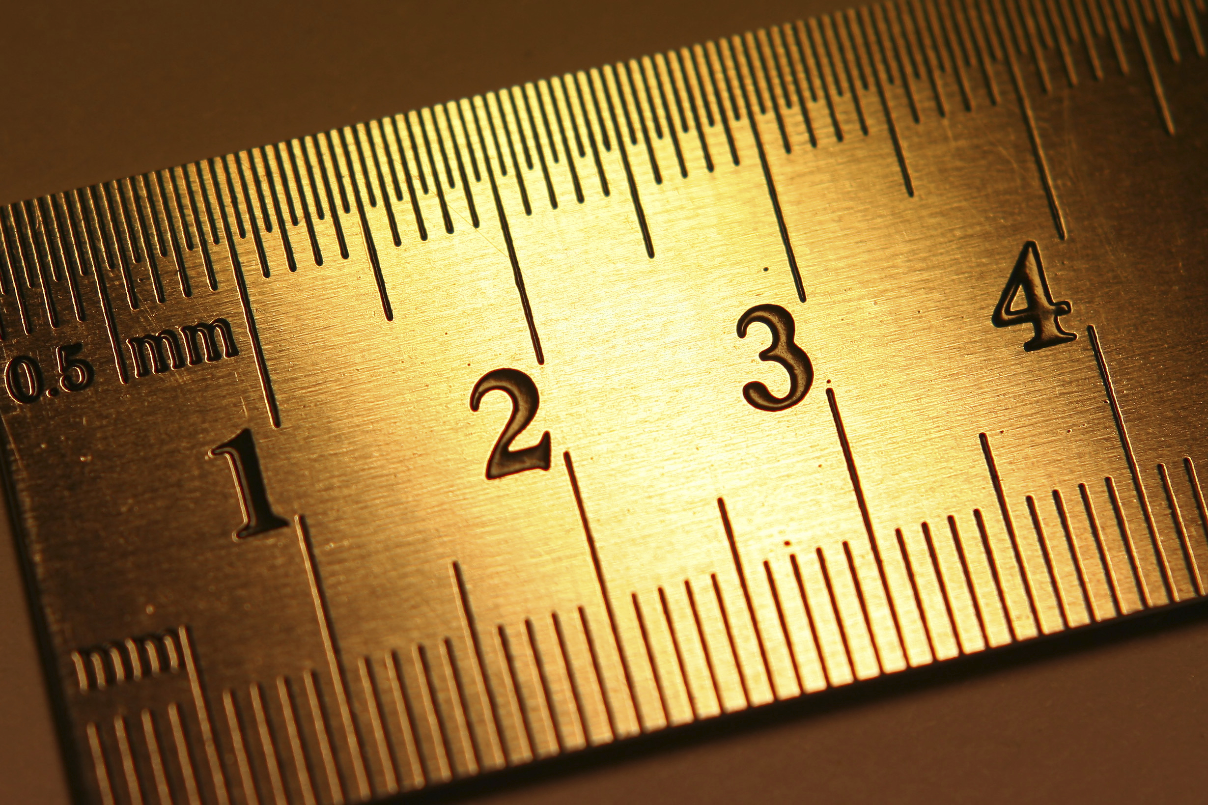 Close-up of a golden metallic ruler with markings for millimeters on one side and numbers 1 to 4 prominently displayed. The lighting highlights the textured surface, creating a warm glow and shadows around the engraved measurements, almost as if illustrating the precise relationships between each mark.