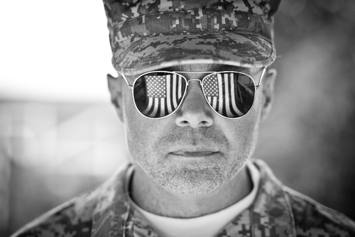 A grayscale image of a soldier wearing a military cap and uniform. He is facing the camera, sunglasses reflecting the American flag in each lens. His expression is neutral, embodying the stoic pride and honor of veterans. The background is blurred, highlighting his resolute presence.