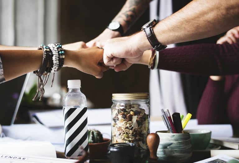 A group of employers joins fists in a gesture of solidarity over a cluttered table with various items such as a jar of mixed nuts, water bottle, and office supplies. All hands display different accessories like bracelets and watches, indicating diverse individuals.
