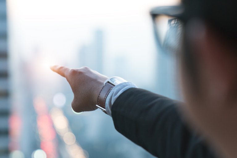 A person wearing glasses and a suit points towards a blurry, sunlit cityscape. The image is taken from behind the person's shoulder, focusing on their extended arm and hand, emphasizing that character matters in navigating the illuminated tall buildings and city lights in the background.