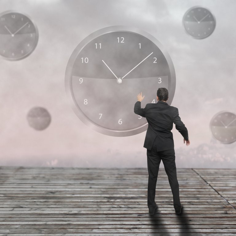A man in a suit stands on a wooden platform, reaching out towards a large clock on a foggy background. Other smaller clocks float around him in the mist, emphasizing themes of time management and the passage of time.