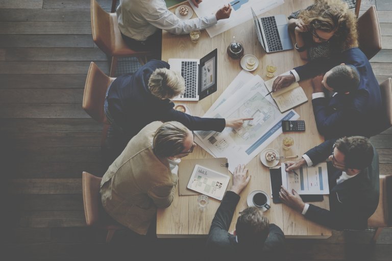 A team of people in business attire sit around a wooden table, engaged in a meeting. Documents, laptops, tablets, coffee cups, and snacks are on the table. One person is pointing at a printed document, and others are looking at laptop screens or holding papers.