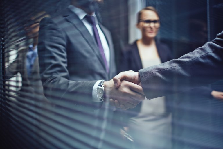 Two businesspeople shaking hands in a professional setting, with blurred figures of other employees in the background. They are dressed in formal business attire, suggesting a corporate environment or a business meeting.