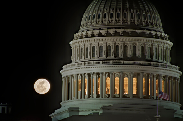 A full moon glows brightly in the night sky beside the illuminated dome of the United States Capitol Building. The architecture features elegant columns and detailed stonework, reminiscent of a grand Federal job interview setting. The American flag is visible in the lower right corner.