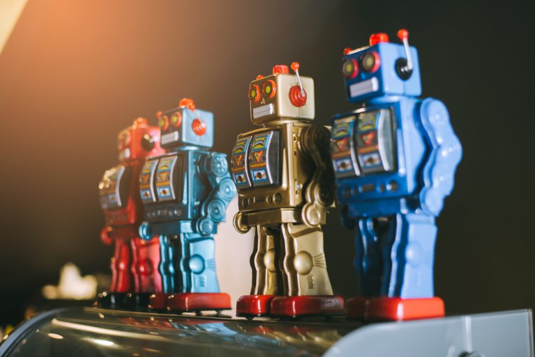 Photograph of three vintage toy robots, hinting at a robot revolution. They are lined up in a row with the first robot in red, the second in silver, and the third in blue. Each robot features colorful details and a square body design, set against a dark background with warm light from the left.