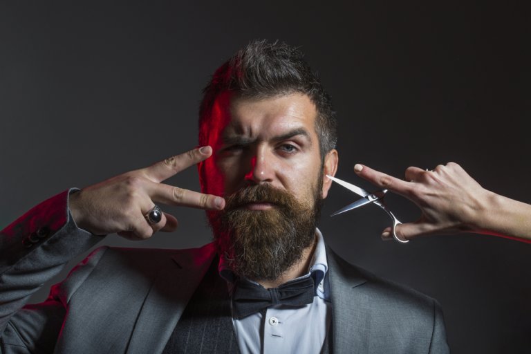 A bearded man in a suit makes peace signs with his fingers, positioning them near his face. A hand holding a pair of scissors is poised near his ear, as if about to cut his hair. The lighting creates a dramatic, almost cinematic atmosphere, elevating the scene into a professional image.