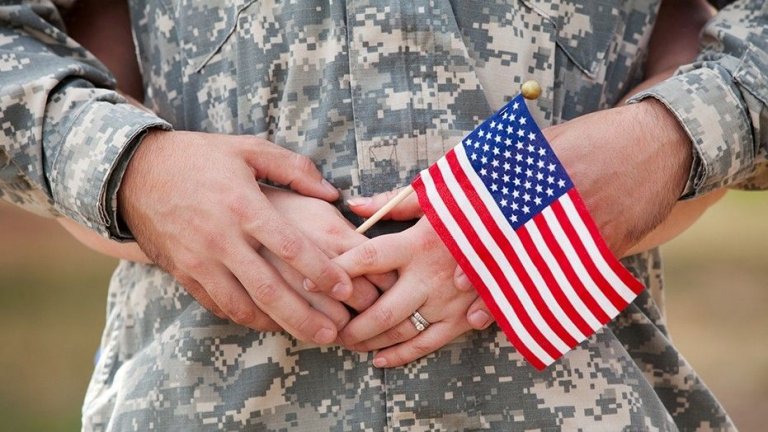 A person in military camouflage uniform holds a small American flag while embracing another from behind. The second person's hands rest on top of the first person's, a ring visible on one finger, symbolizing the deep bond shared by military spouses.