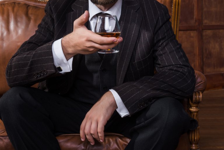 A well-dressed man in a pinstripe suit sits on a leather chair, holding a glass of cognac. His face is partially obscured, directing attention to his attire and the drink. The background features wooden paneling, subtly hinting at a room where teamwork and sophistication seamlessly blend.