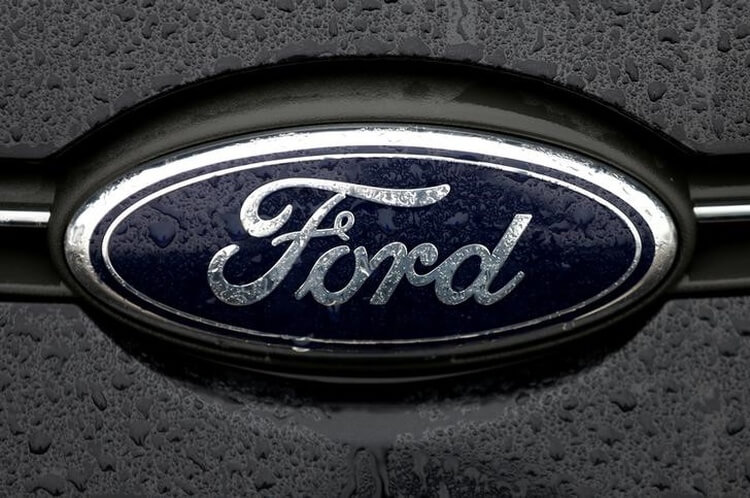 Close-up photograph of a Ford emblem on a car, displaying the iconic blue oval with the word "Ford" in cursive white letters. The surface is wet with raindrops, indicating recent rainfall in Detroit, Michigan. The background is slightly blurred and dark.