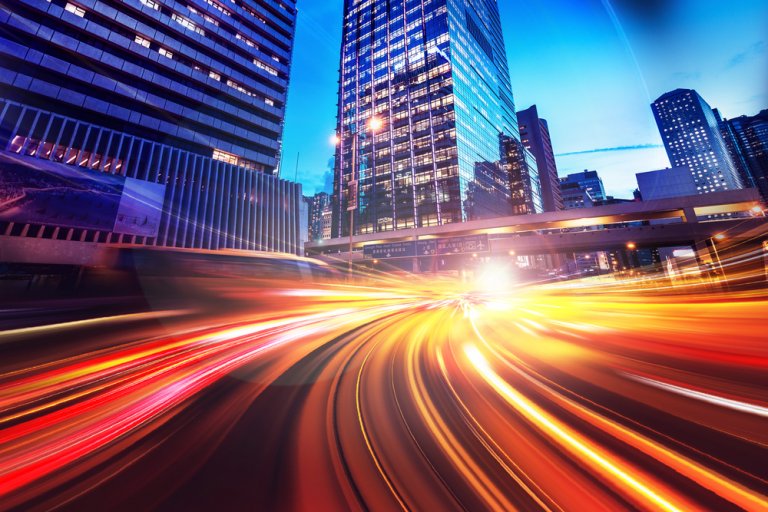 A vibrant cityscape at night featuring tall skyscrapers, much like the meticulous crafting of a cover letter. The image showcases light streaks from fast-moving vehicles on a road, creating dynamic lines of red, yellow, and white light against the bright lights of the city buildings.
