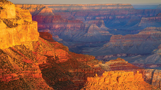 A stunning view of the Grand Canyon during sunset showcases the vast expanse of the canyon with its layered reddish-orange rock formations and deep valleys. The light from the setting sun casts a warm glow, highlighting the rugged terrain that plays a crucial role in driving Arizona's economy through tourism.