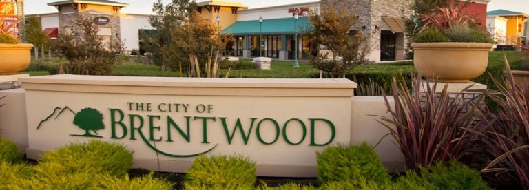 A landscaped sign reads "The City of Brentwood" with greenery and decorative plants in the foreground. Behind the sign, various facades of buildings and shops create a lively shopping area, adding charm to Brentwood's inviting town center.
