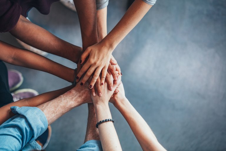 A diverse group of people stands in a circle, stacking their hands together in the center to symbolize unity, teamwork, and engagement. The background is a neutral gray, and the image is taken from a top-down perspective.