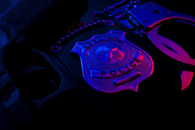 A close-up photograph of a "Special Police" badge, a pair of handcuffs, and a firearm, illuminated by blue and pink lighting against a dark background. The police badge is central, with the chain, handcuffs, and gun partially visible.