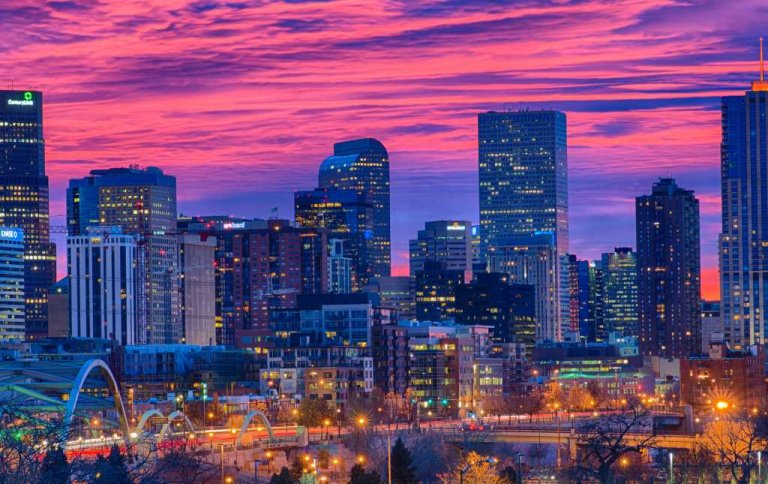 A vibrant Denver cityscape at dusk showcases a variety of tall buildings with lights starting to turn on. The sky is painted in shades of pink, purple, and orange, creating a stunning sunset backdrop. The foreground features a bridge with illuminated streetlights.