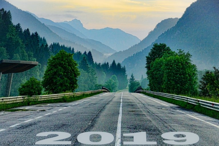 A paved road with "2019" painted at the bottom stretches into a misty landscape. Flanked by lush green trees and mountains fading into the distance under a serene, cloudy sky, the scene evokes a sense of calm and progression.