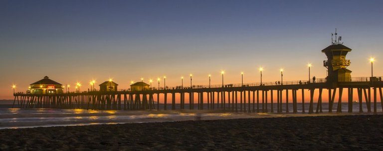 A long, illuminated pier stretches into the ocean against a twilight sky, with calm waves lapping at the shore and the horizon painted in hues of orange and blue. Lights on the pier and buildings add a warm glow to the peaceful scene.
