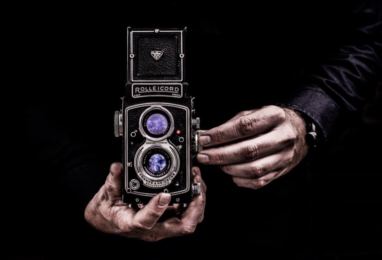 Close-up of a person holding a vintage Rolleicord Twin Lens Reflex camera against a dark background. The person's hands are adjusting the camera, highlighting its intricate details and reflective lenses. The scene is dramatically lit, emphasizing the vintage experience and aesthetic.