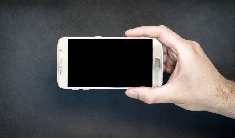 A person holding a smartphone horizontally with a black screen, perfect for scrolling through social media. The phone has a metallic frame and a prominent home button. Against a simple dark gradient background, the hand is positioned to showcase the screen.