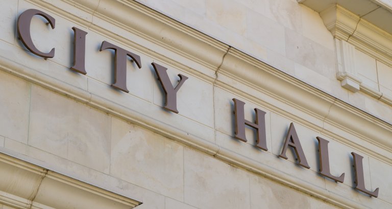 Close-up of the exterior of a stone building with the words "City Hall" prominently displayed in large, dark letters. The light beige facade enhances the building's brand, with the letters mounted above an architectural molding detail.