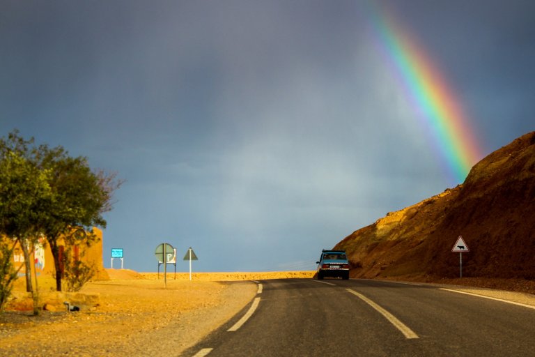 A car drives on a two-lane paved road in an arid landscape with rocky hills. A vibrant rainbow arcs across a dark, cloudy sky, symbolizing the journey toward increased employee engagement. Road signs are visible on the side of the road, and a small building is partially seen in the distance.