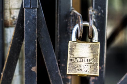 Close-up image of a brass padlock labeled "ABUS Patented N-83/45" securing a metal gate. The gate is made of rusted metal bars overlapping at various angles, contributing to an industrial and weathered appearance, symbolizing strong cybersecurity measures. The focus is sharp on the padlock and surrounding gate area.
