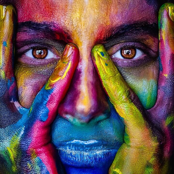 A close-up of a person with hands framing their face, featuring vivid and colorful face paint. The paint covers the face and hands in bright hues—red, blue, green, and yellow—creating a striking artistic impression; the eyes prominently visible seem to ask, "What do you know?