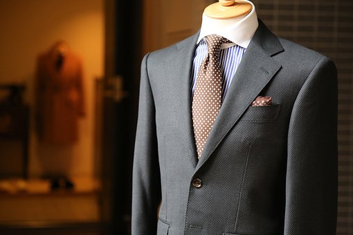 A close-up of a mannequin dressed in a dark gray suit with a light blue striped shirt and a brown polka-dot tie, perfect for making an impression at your next cover letter interview. A brown coat hangs in the blurred background, all displayed in a well-lit store.