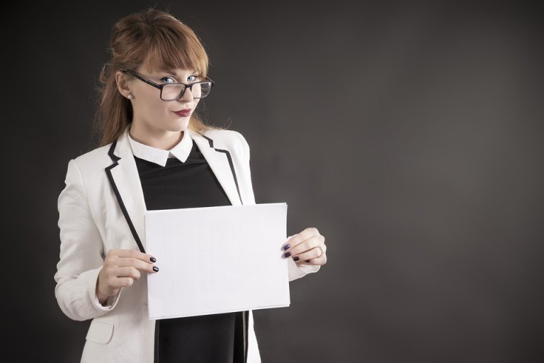 A woman with glasses and a white blazer over a black top holds a blank piece of paper marked "ATS" in front of her. She is looking slightly to the side with a neutral expression against a dark background.