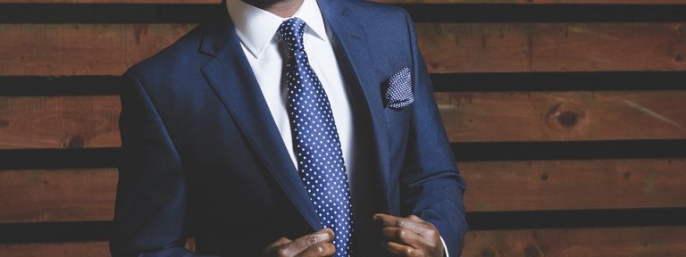 A confident candidate stands against a wooden background, dressed in a dark blue suit, white dress shirt, and blue polka dot tie. He adjusts his suit jacket with both hands, exuding professionalism. A patterned pocket square is neatly folded in his breast pocket.