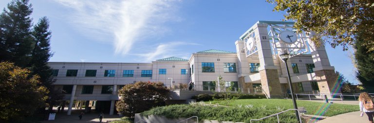 Panoramic view of Sonoma State's modern, multi-story academic building with large windows and green-tinted roofs. The building is surrounded by trees and well-manicured lawns, with a clock mounted on a pole in the foreground and a person walking down a path on the right.