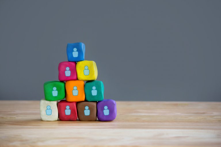 A pyramid of colorful clay blocks sits on a wooden surface against a gray background. Each block displays a white icon of a person, symbolizing unity during COVID-19. The pyramid consists of ten blocks in various colors such as blue, yellow, green, red, orange, purple, brown, and teal.