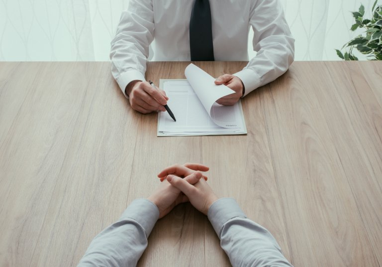 Two people seated across a wooden table from each other, discussing matters amid the COVID-19 pandemic. One, visible from the waist up in a white shirt and tie, holds a pen and papers. The other, with hands clasped, is only partially visible from the arms down.