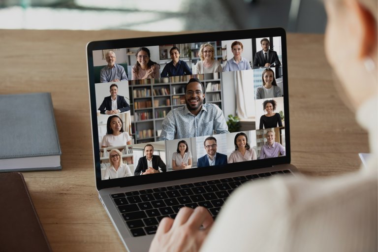 A person is participating in a video conference on a laptop, showing 12 small screens of diverse individuals in a grid format. The participants are engaging, smiling, and appear to be discussing COVID-19 staffing solutions. There are bookshelves and home environments in the background.