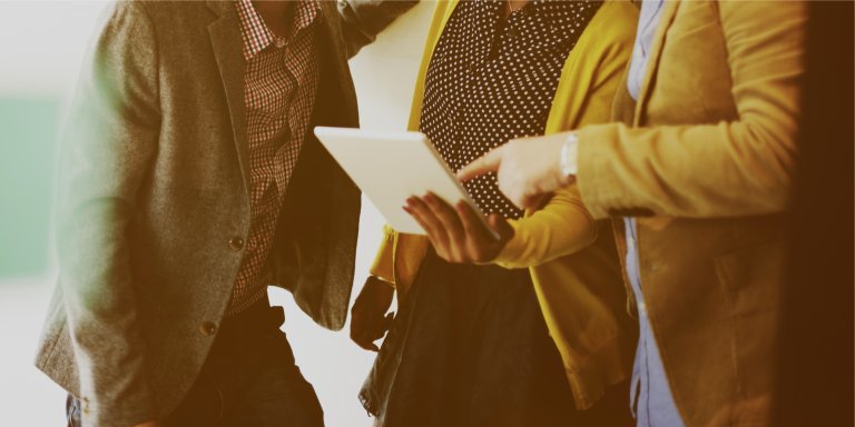Three people in business casual attire are gathered closely, viewing and discussing something on a tablet. Their faces are not visible, with focus on the tablet and their hand gestures. They appear to be collaborating in an indoor setting, likely working on an HR project.